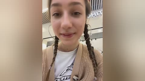 Media: Video of a young woman with light skin and braided hair, wearing a beige cardigan over a white graphic t-shirt, smiling in a modern indoor setting with white ceiling tiles and a partial \"THE\" sign in the background.