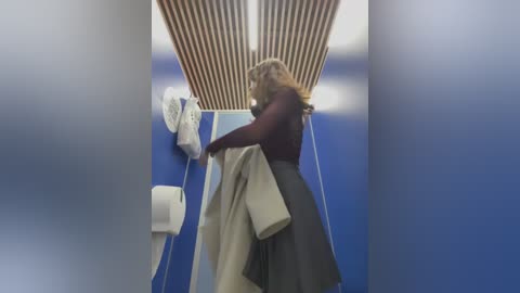 Media: Video of a woman with curly hair, wearing a maroon top and grey skirt, hanging coats in a narrow, blue-walled hallway with wooden slats on the ceiling and a toilet paper dispenser on the left wall.