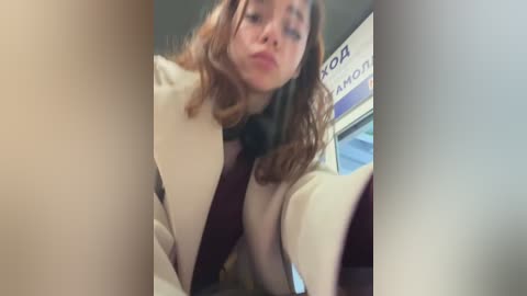 Media: A video of a young woman with shoulder-length wavy hair, wearing a beige coat and black scarf, taken from a low angle, capturing a casual, candid moment indoors.