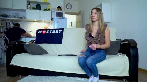 Media: Video of a blonde woman in a sleeveless top and jeans playing a video game on a white couch in a modern kitchen.