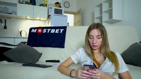 Media: Video of a blonde woman with fair skin, small breasts, wearing a white blouse, sitting on a beige couch, reading a book, with a remote control and a blue MSTBET pillow behind her.