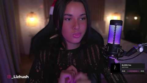 Media: Video of a Latina woman with long black hair, wearing a black sheer top, recording a live stream on Twitch. The background shows a dimly lit room with wooden furniture and soft lighting.