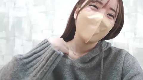 Media: Video of a young Asian woman with straight, shoulder-length dark brown hair, wearing a gray sweater and a beige surgical mask, holding her shoulder. The background is blurred, showing a light-colored room.