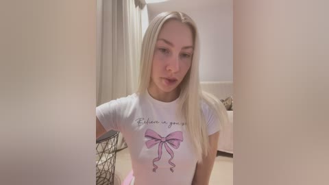 Media: Video of a blonde woman with fair skin, wearing a white t-shirt with a pink bow graphic, posing indoors with beige walls and furniture.