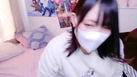 Media: Video of an Asian woman with long black hair wearing a white mask, a white shirt, and a choker, standing in a bedroom with a blue-patterned pillow, anime posters, and a pink bedspread.