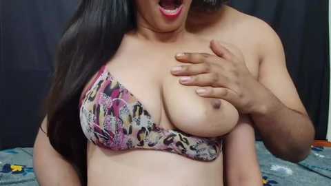 Media: Video of a light-skinned woman with long black hair, wearing a colorful bra, exposing her large breasts, being grabbed from behind by a darker-skinned person, in a dimly lit room.