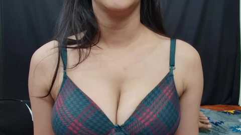 Media: Video of a light-skinned woman with long black hair, wearing a low-cut, green and purple plaid bra, her cleavage prominently displayed against a dark, textured background.