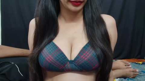 Media: A video of a South Asian woman with long black hair, fair skin, and red lipstick, wearing a dark blue plaid bra with pink accents, seated in a room with a black curtain backdrop and a partially visible person's feet.