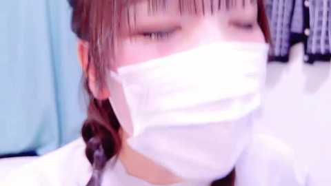 Media: Video of a woman with light skin and brown hair, wearing a white surgical mask, lying on a bed in a hospital setting. The background includes a light blue curtain and a blurry medical device.