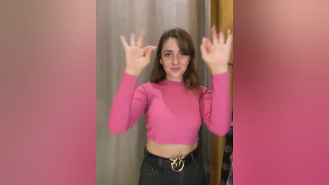 Media: Video of a young woman with fair skin, wavy brown hair, and wearing a pink crop top and black pants, making a peace sign in front of a pink and beige curtain backdrop.