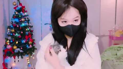 Media: Video of an Asian woman with long black hair, wearing a black mask, holding a white rabbit, in a festive room with a decorated Christmas tree and a pink shelf.