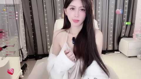 Media: Video of a slender, East Asian woman with long, straight black hair, wearing a white off-shoulder top, sitting in a modern office with grey curtains and a white desk.