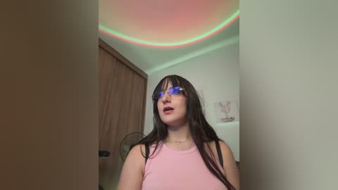 Media: Video of a young woman with long, straight dark hair, wearing blue sunglasses, pink tank top, and looking up. Background includes a wooden door, a ceiling with green and pink lights, and a white wall.