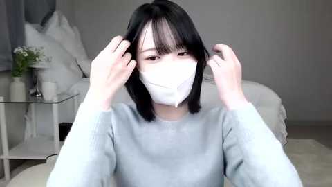 Media: Video of a young Asian woman with straight black hair, wearing a light gray sweater and white surgical mask, adjusting her hair, in a minimalist bedroom with white furniture and a bed.