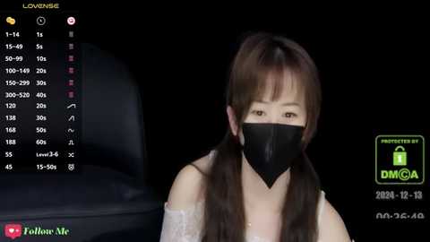 Media: Video of a young Asian woman with long brown hair, wearing a black face mask, sitting indoors. The background is dark, and the image includes a black leather couch and a computer screen displaying a live stream.