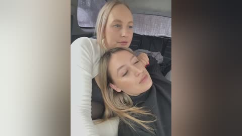 Media: Video of two young women, one blonde with a white top, the other brunette with a black top, lying on a bed with a grey comforter.