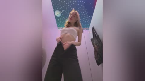 Media: Video of a young, slender woman with blonde hair, wearing a white bandeau top and black pants, standing in a narrow, white hallway with a celestial mural on the ceiling.