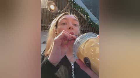 Media: Video of a blonde woman with fair skin, wearing a black sweater, drinking from a yellow cup. She's in a dimly lit, outdoor wooden setting with string lights and a bamboo fence in the background.