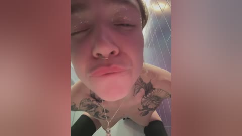 Media: Video of a young woman with short hair, nose ring, and tattoos on her shoulders, sitting in a bathroom with blue tiles and a showerhead in the background.