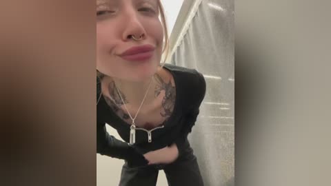 Media: Video of a young woman with light skin, pouting, wearing a black zip-up jacket and black pants, with tattoos visible on her chest. She stands in a shower with a white curtain in the background.