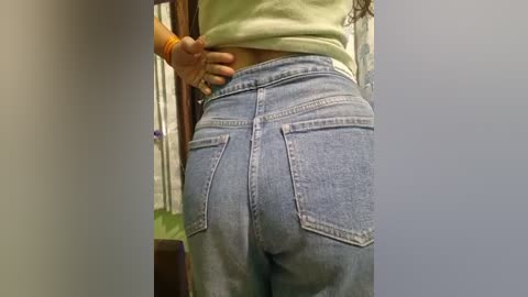 Media: Video of a person from the back, wearing light blue denim jeans with a large back pocket. The person is partially visible in a green shirt. The background shows a beige wall and a wooden door frame.