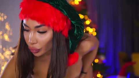 Media: Video of a Latina woman with long brown hair, wearing a green velvet Santa hat with a red pom-pom, adjusting it, in a festive indoor setting with bokeh lights and a Christmas tree.