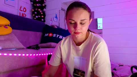 Media: Video of a young girl with light skin, brown hair, wearing headphones, seated on a beige couch in a dimly lit room with purple LED lights, a Christmas tree, and \"LOST\" pillow.