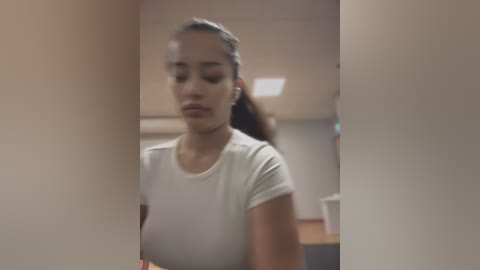 Media: Video of a young woman with light brown skin and medium-length dark hair, wearing a tight white top, standing in a dimly lit room with a blurred background.