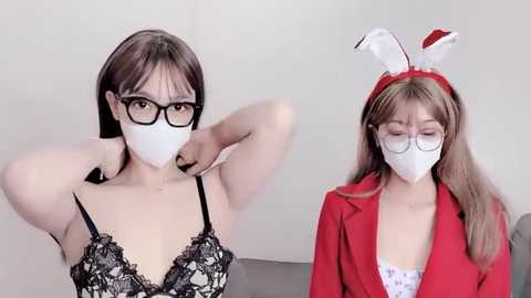 Media: Video of two women in face masks, one in black lace lingerie, the other in a red blazer and bunny ears, both indoors with minimal background.