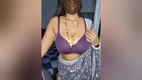 Media: Video of a woman with medium skin tone, wearing a purple bra, gold necklace, and gray floral-patterned sari, standing in a room with a checkered patterned blanket and brown curtains.