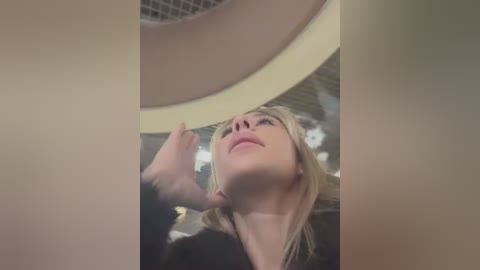 Media: Video of a blonde woman with pale skin, wearing a black top, standing inside a cylindrical structure, with her head tilted back, looking upward.