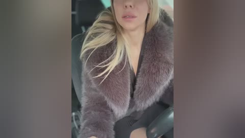 Media: Video of a blonde woman in a grey fur coat, sitting in a car, with a blurred background.