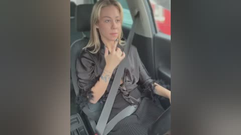 Media: Video of a blonde woman in a black shirt, wearing a seatbelt, seated in a car with a red object visible outside.