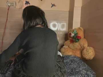 Media: A video of a person with shoulder-length black hair wearing a dark sweater, sitting on a bed with a teddy bear and a patterned blanket. The room has beige walls with decals and a fan on the wall.