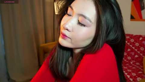 Media: Video of an Asian woman with long black hair, wearing a red top, sitting in a room with beige curtains and a red floral couch. She has a serene expression with eyes closed, suggesting relaxation or meditation.