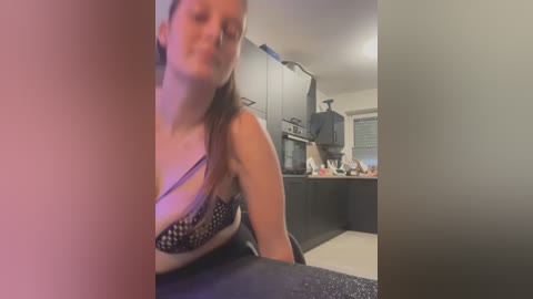 Media: Video of a young woman with light skin and long brown hair, wearing a black and white patterned bra, sitting in a modern kitchen.