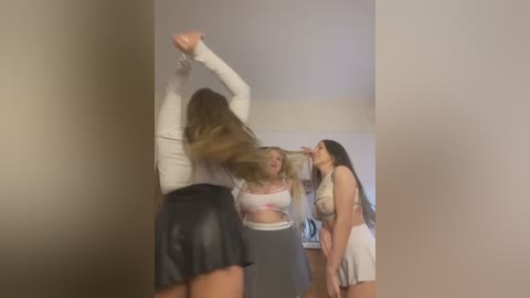 Media: Video of three young women dancing energetically in a dimly lit room. Two wear tight, revealing outfits; one in a white crop top and black skirt, another in a white crop top and high-waisted shorts. Background features a plain, beige wall.