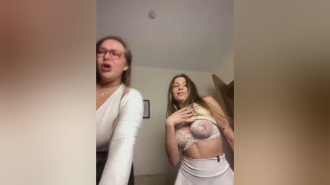 Media: Video of a blonde woman in glasses and a white top, and a brunette in a white lace bra and skirt, both looking surprised in a dimly lit room.