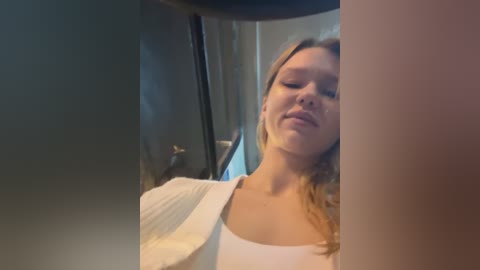 Media: Video of a young woman with fair skin and long blonde hair, wearing a white shirt, standing in a dimly lit room, partially reflected in a mirror.