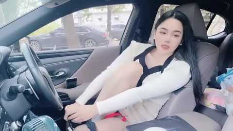 Media: Video of a young East Asian woman with long black hair, wearing a white cardigan, sitting in a car seat with a black seatbelt. Background shows a car parked outside with a blurry tree and parked vehicles visible through the window.