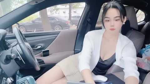 Media: Video of an East Asian woman with fair skin, dark hair in a bun, wearing a white blazer over a black top and beige skirt, sitting in a car with beige seats, looking relaxed.