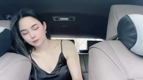 Media: Video of an Asian woman with long black hair, fair skin, and wearing a black satin camisole, sitting in the front seat of a car with beige upholstery, eyes closed, appearing relaxed.