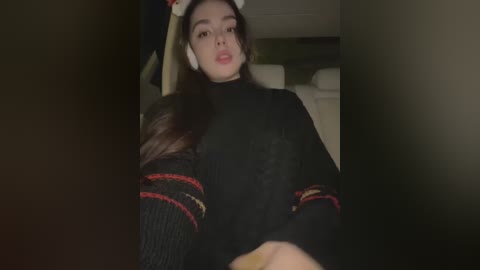 Media: Video of a young woman with long, dark hair, wearing a black sweater with red and gold stripes, sitting in a dimly lit room.