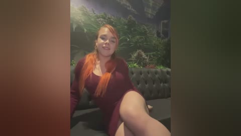 Media: Video of a curvy, fair-skinned woman with long, orange hair, wearing a maroon dress, sitting on a tufted black leather couch in a dimly lit room with lush greenery in the background.