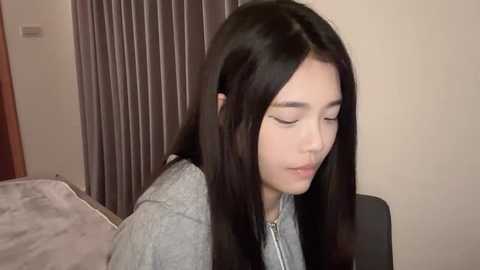 Media: Video of a young Asian woman with long, straight black hair, wearing a light gray hoodie, sitting indoors with closed eyes and serene expression. Background includes beige walls and vertical brown curtains.