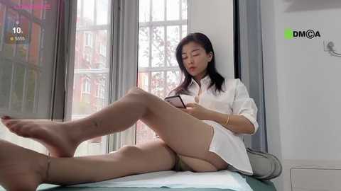 Media: A video of an Asian woman with long black hair, dressed in a white blouse and beige stockings, sitting on a bed, reading a book, in a modern room with large windows and bookshelves.