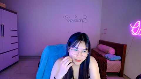 Media: A video of an East Asian woman with long black hair, wearing glasses and a black top, sitting in a room with a blue blanket, a white dresser, a bed with pink pillows, and a neon sign.