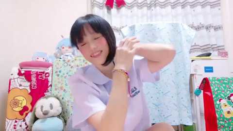 Media: Video of a smiling Asian woman in a light blue shirt, adjusting her shirt, surrounded by stuffed animals and colorful decorations.