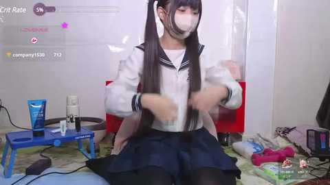 Media: Video of an East Asian woman with long black hair, wearing a school uniform, sitting on the floor in a cluttered room.