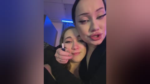 Media: Video of two young women, one with light skin and dark hair, the other with fair skin and blonde hair, making kissy faces while holding a selfie stick in a dimly lit room.